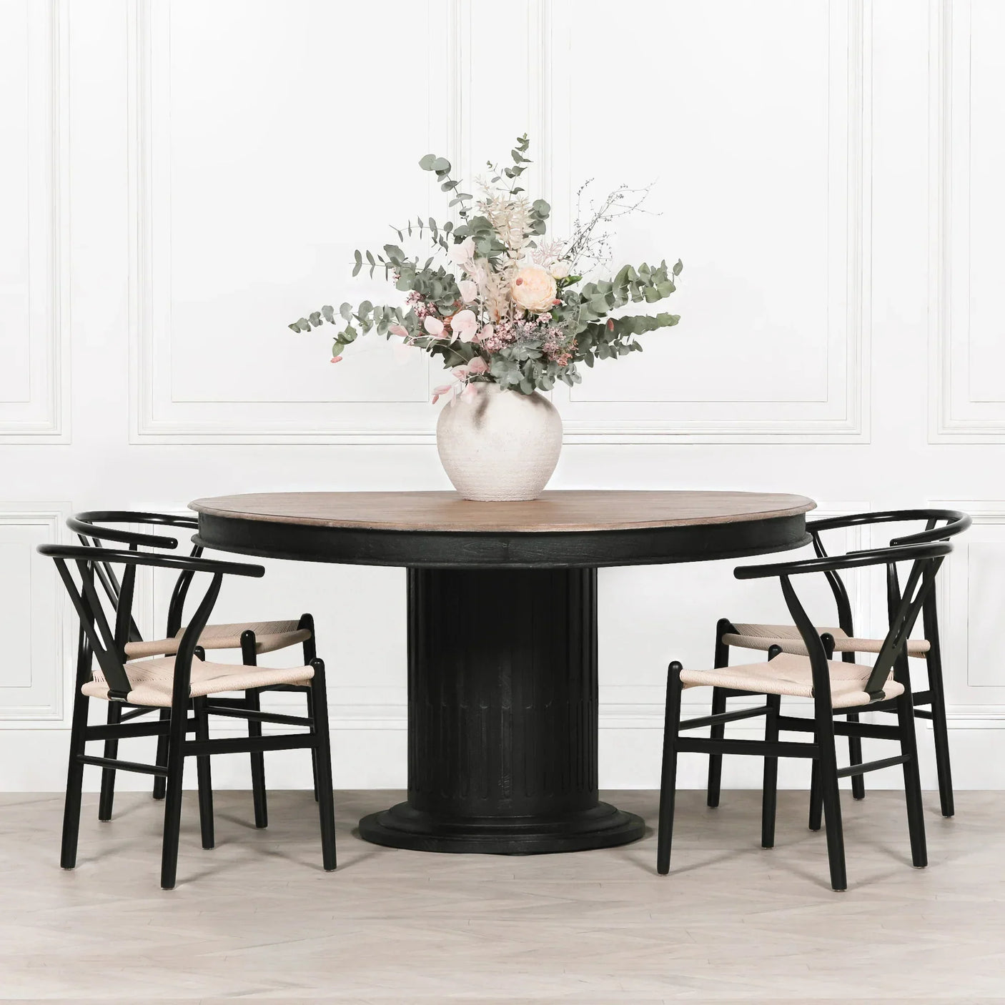 Round black dining table with light wood top and three black chairs, floral vase centerpiece