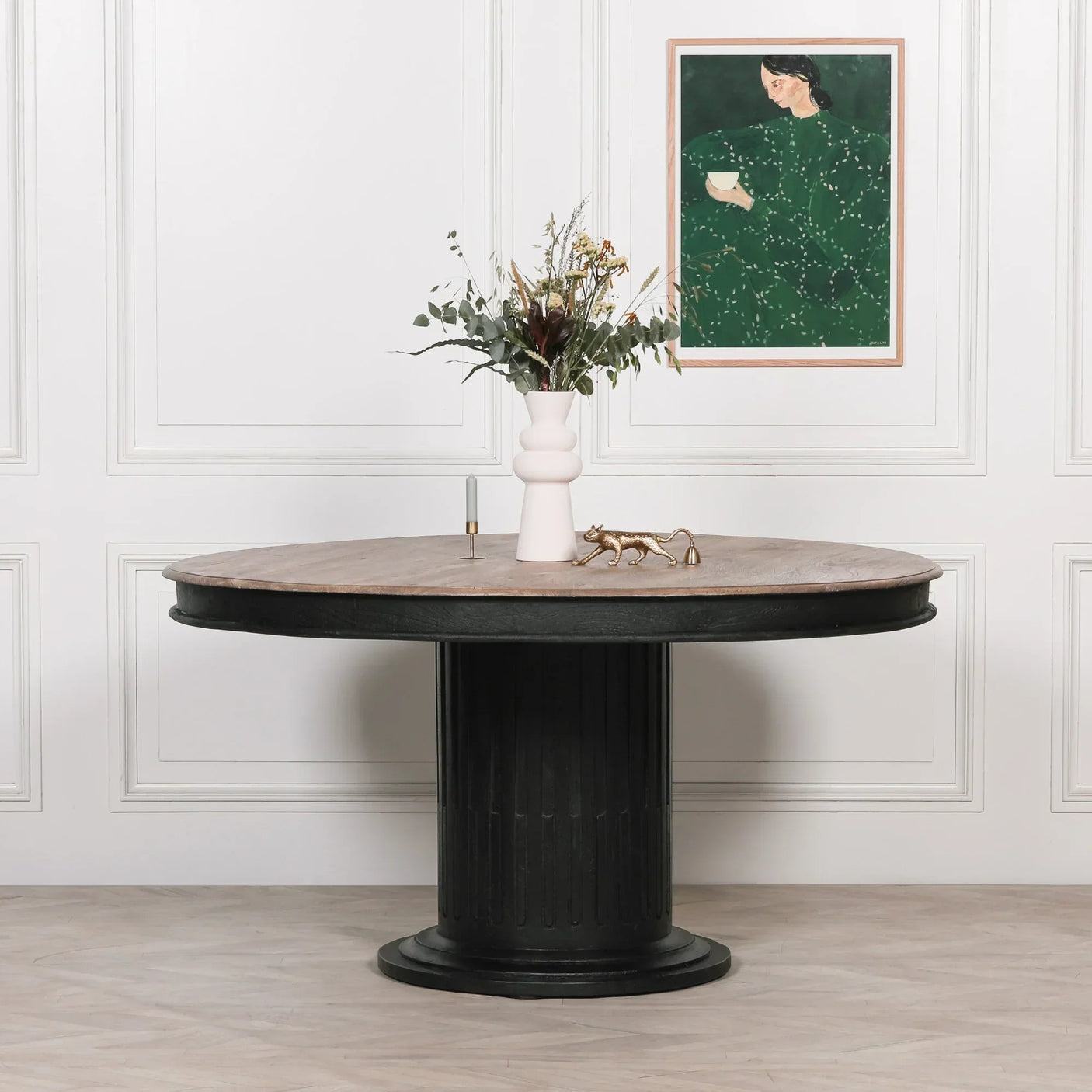 Round dining table with light wood top and black pedestal base, vase and decor on top