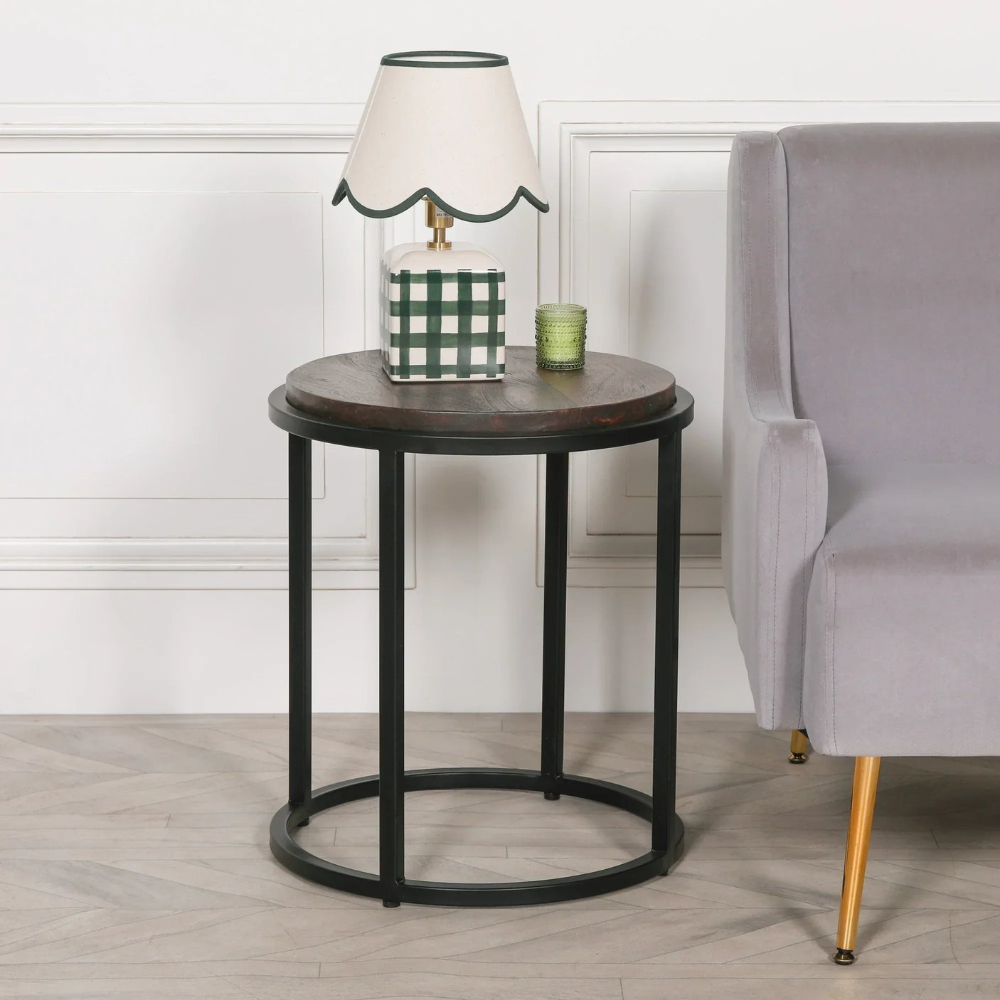 Round wooden side table with black metal frame beside grey armchair and table lamp