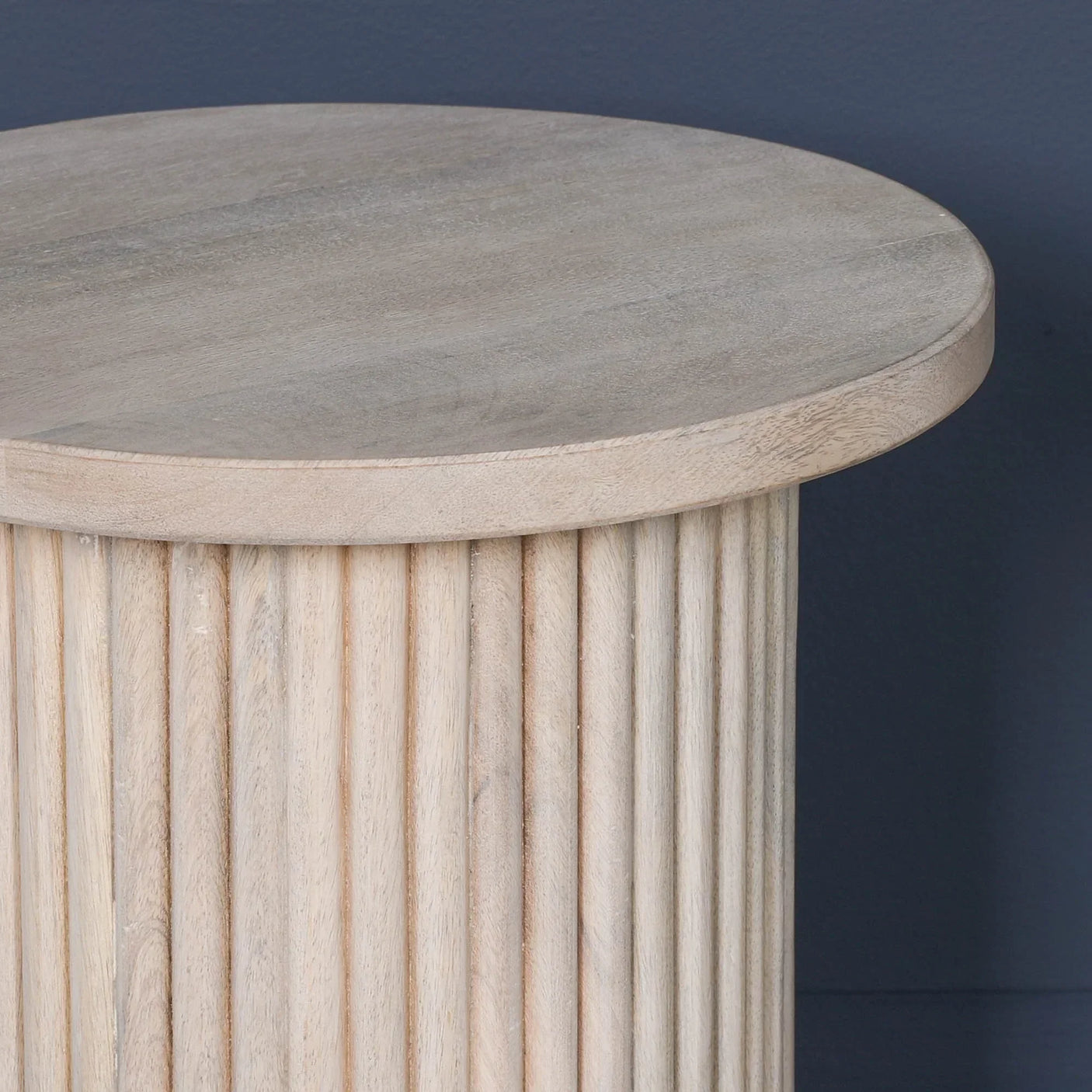 Round wooden side table with ribbed base and light natural finish against dark wall