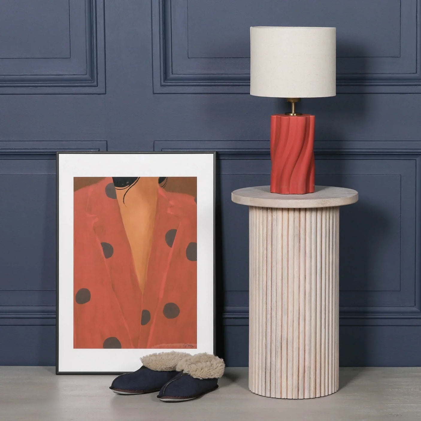 Fluted wood side table with red ceramic lamp, framed art and slippers on floor beside