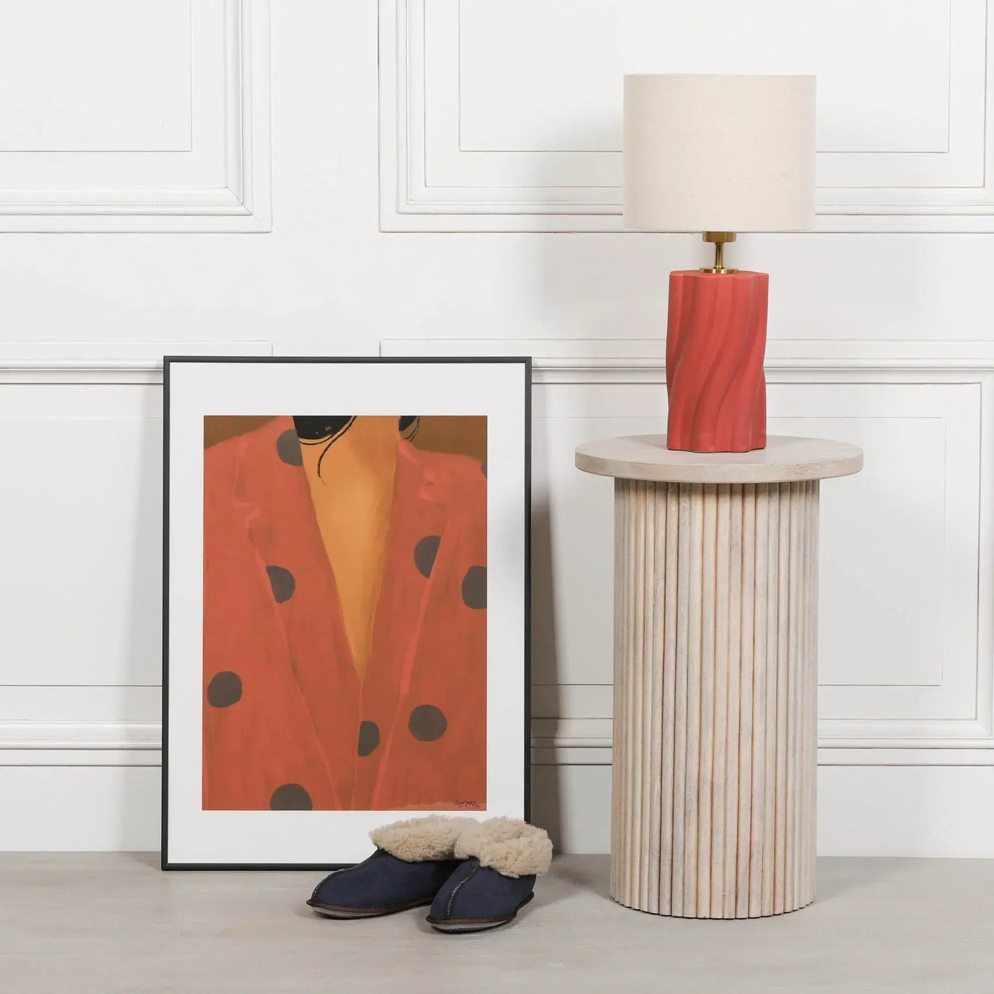 Wooden side table with fluted detail, red ceramic lamp, framed wall art, and slippers
