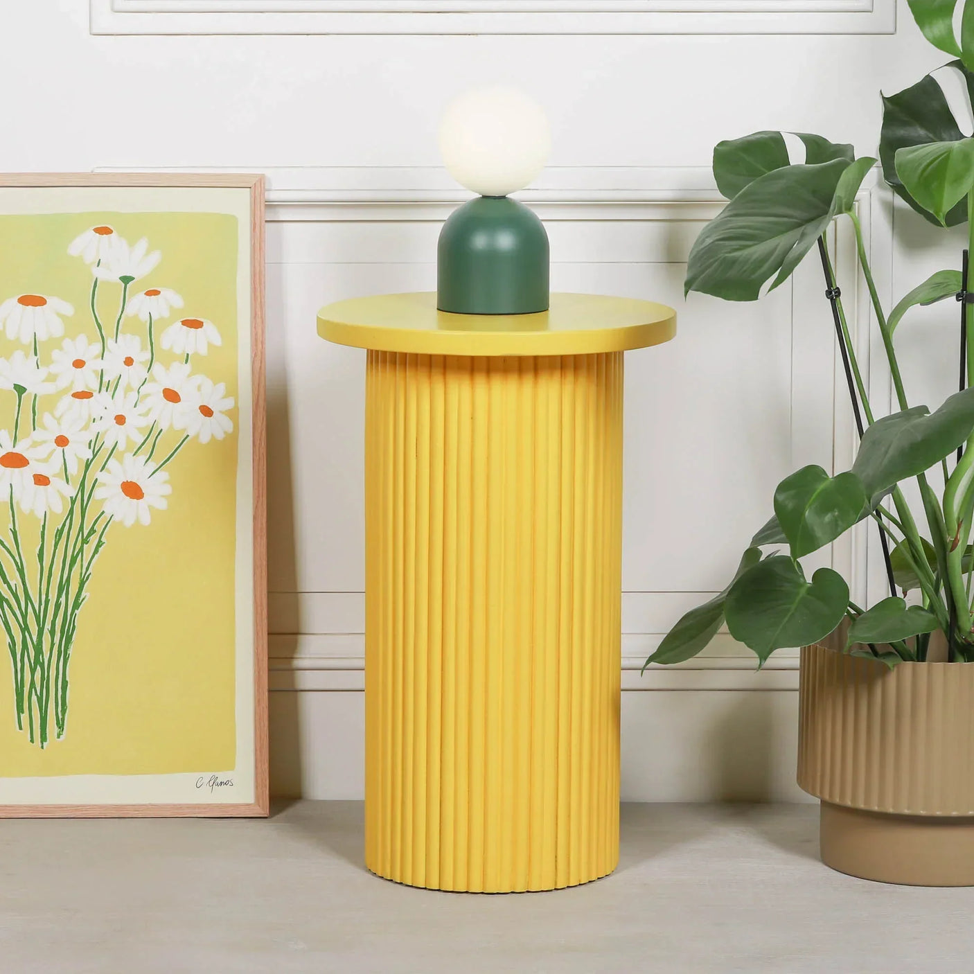 Yellow ribbed side table with round top, lamp, artwork and potted plant beside