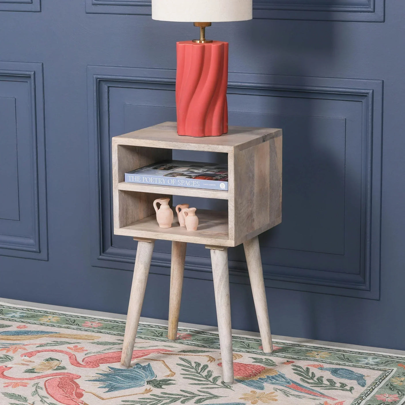 Wooden bedside table with open shelf, lamp, books, and ceramic vases against blue wall