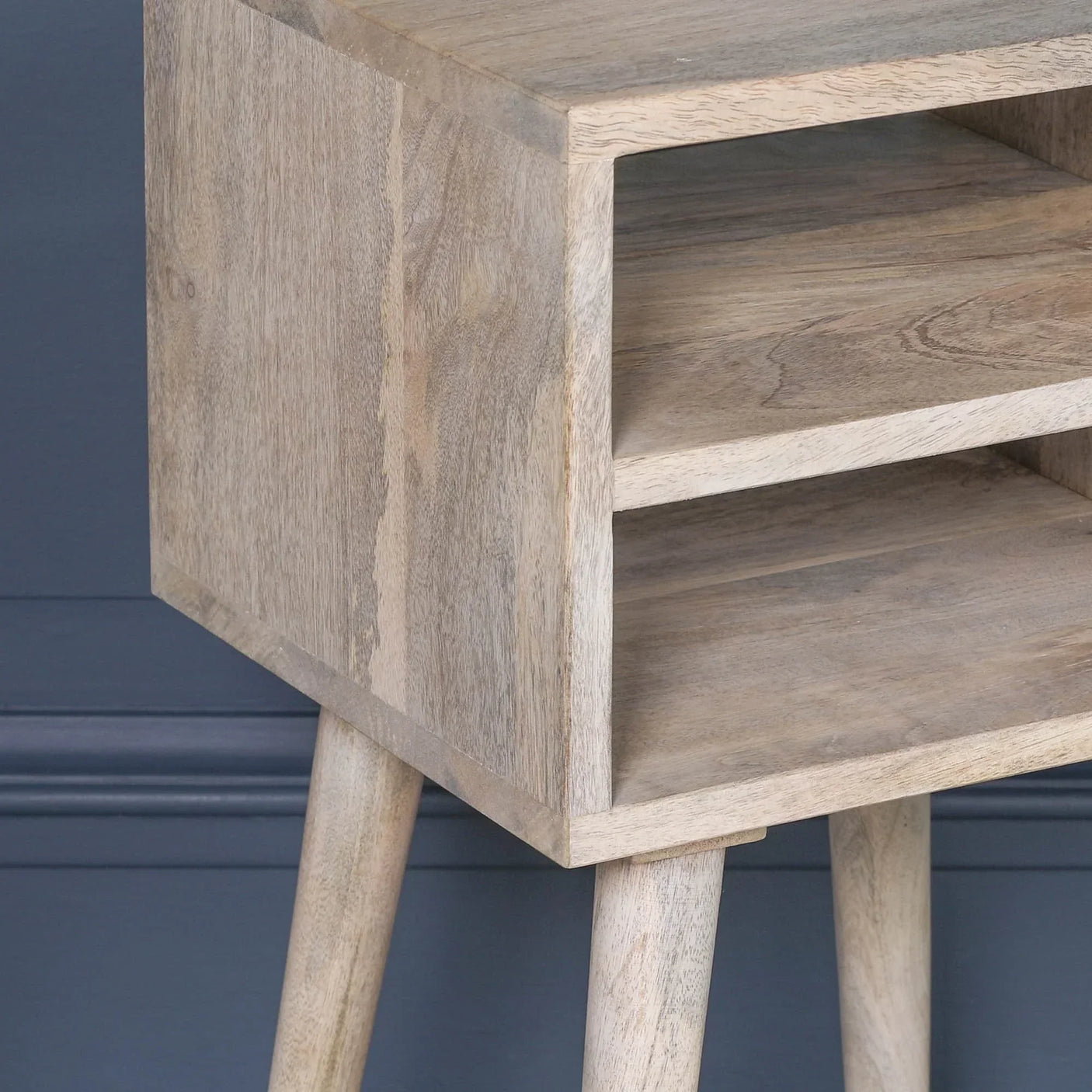 Natural wood side table with angled legs and open storage shelves against blue wall