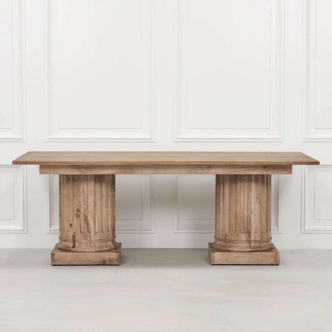 Rectangular wooden dining table with two fluted pedestal bases against white panel wall