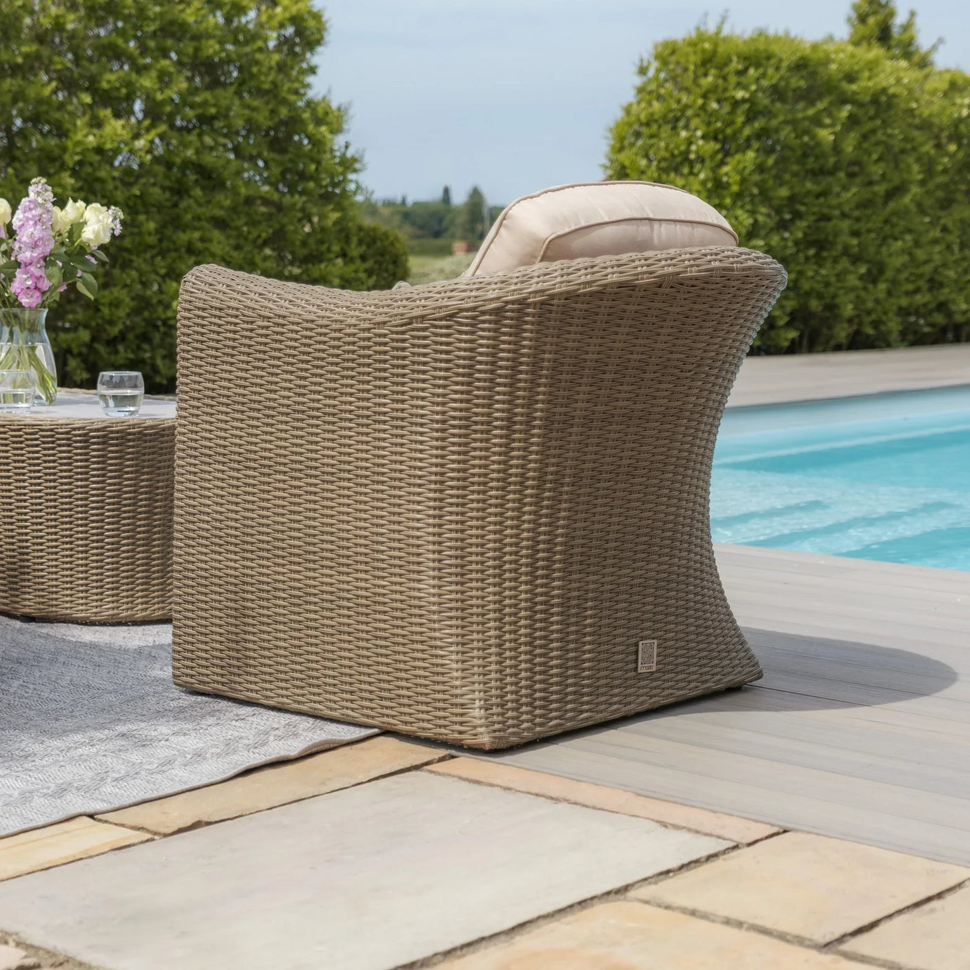 Rattan garden armchair with cushion by poolside on patio with greenery in background