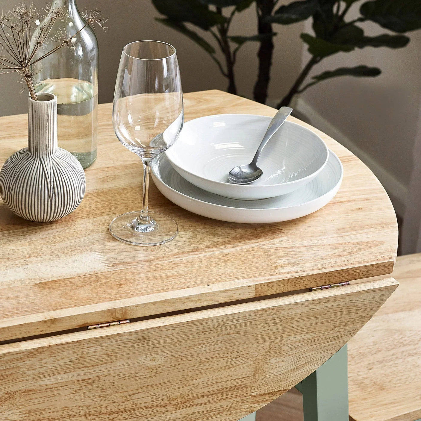 Light wood dining table with drop leaf detail, set with plates, glass and vase