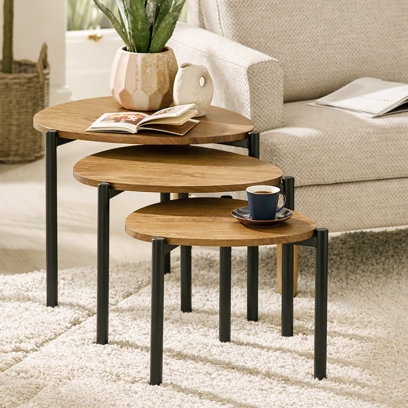Three nesting side tables with light wood tops and black legs in living room with white armchair.