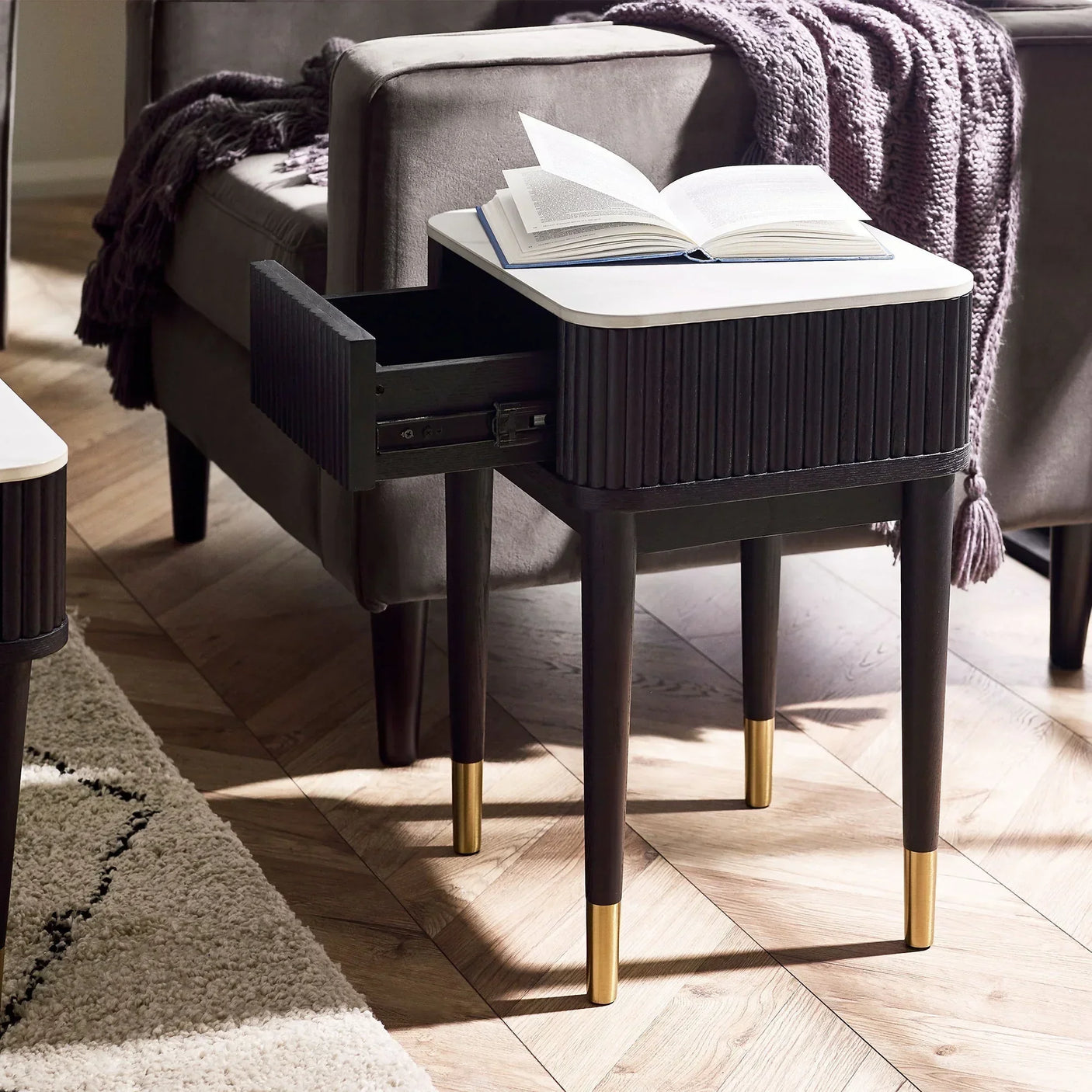 Black fluted lamp table with white stone top and open drawer beside grey sofa in living room