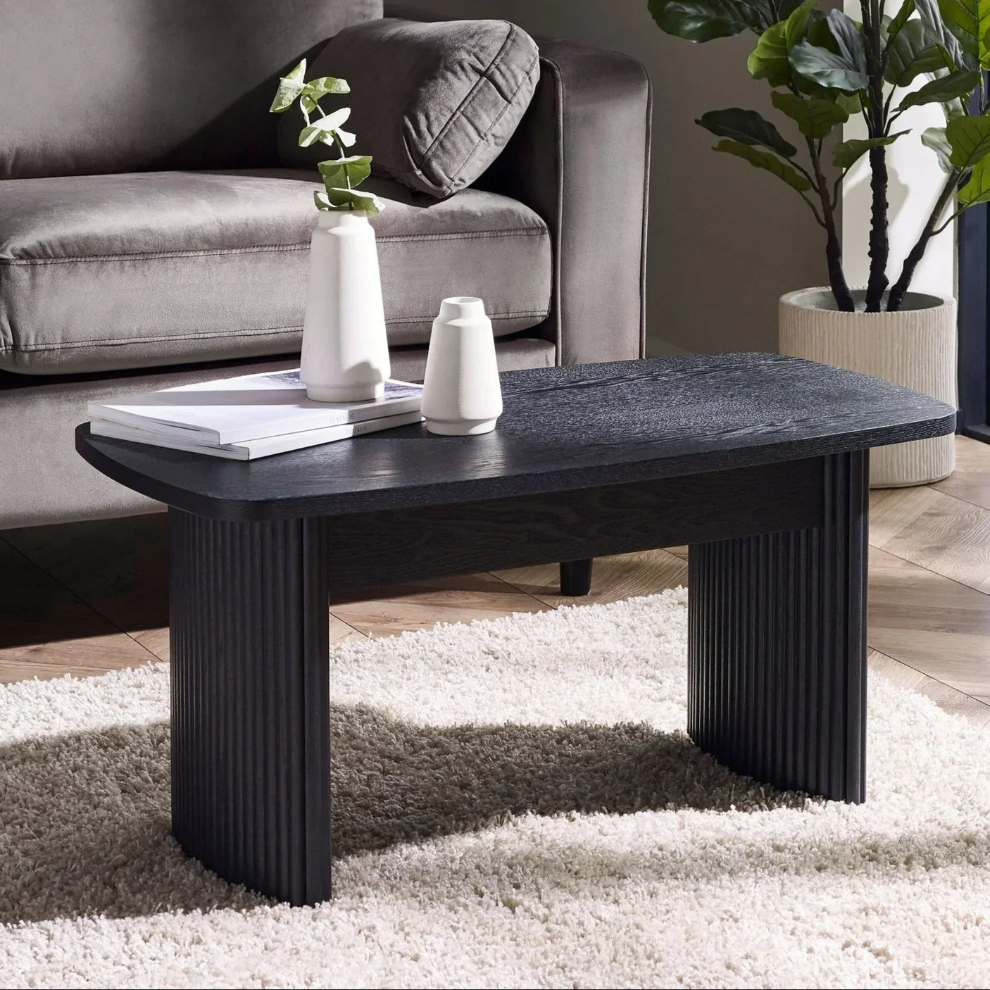 Black fluted compact coffee table in living room with grey sofa, white vases and rug