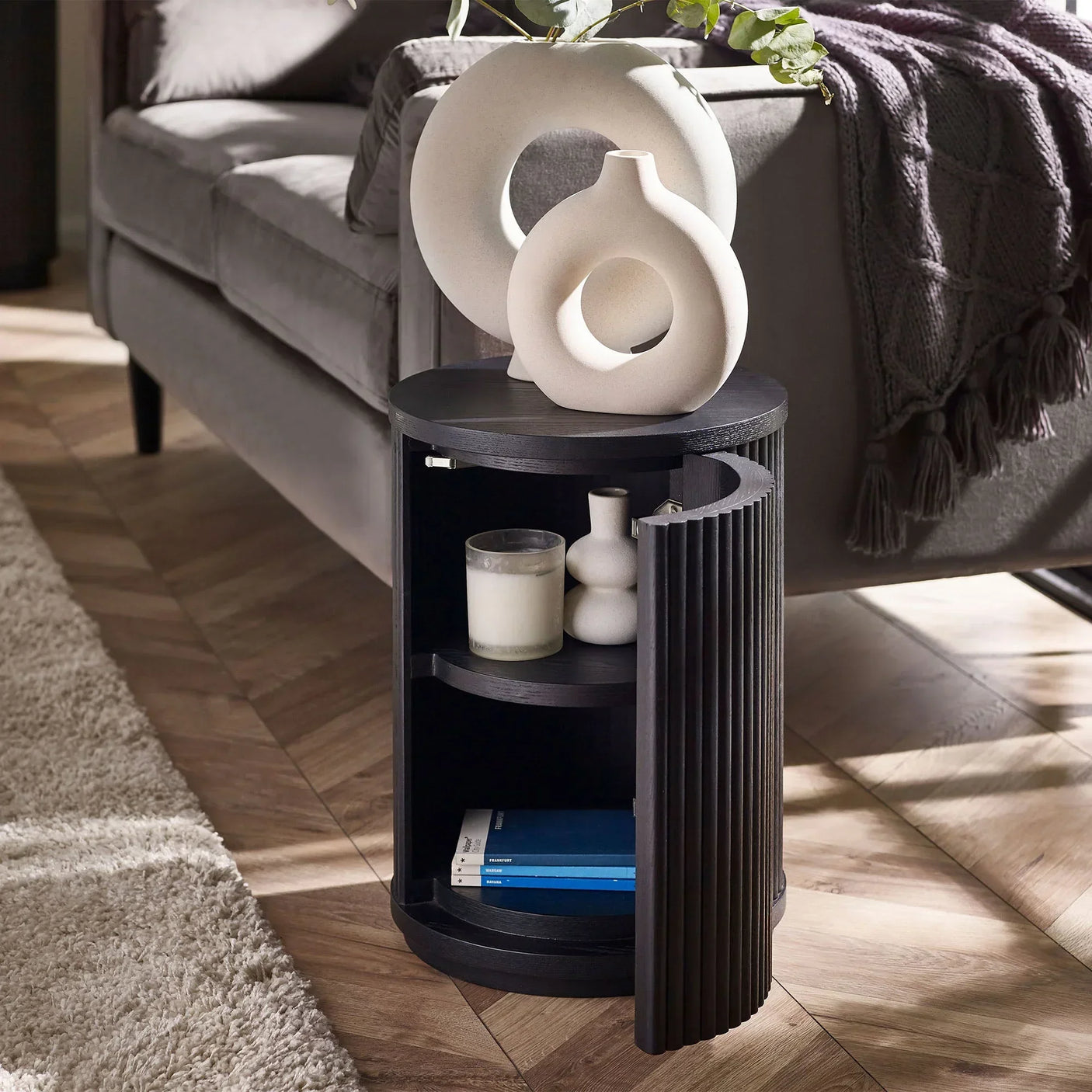 Black fluted side table with door open, shelves holding books and decor, beside grey sofa