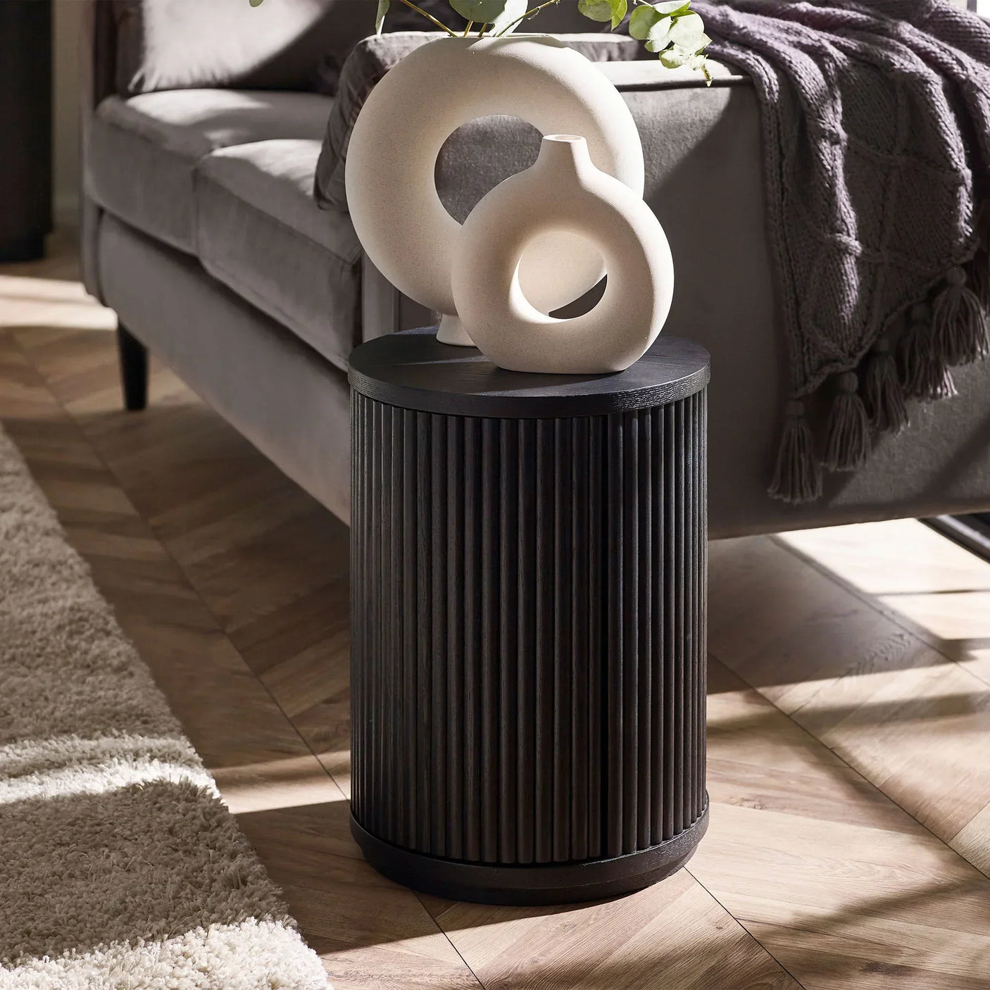 Black fluted side table with door beside grey sofa, styled with ceramic vases