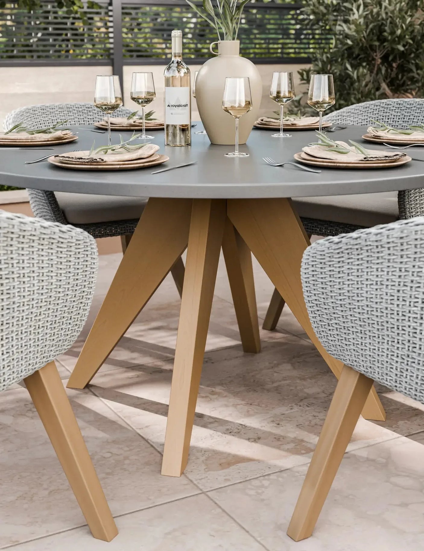 Round outdoor dining table with grey top, wooden legs, and woven armchairs set for four