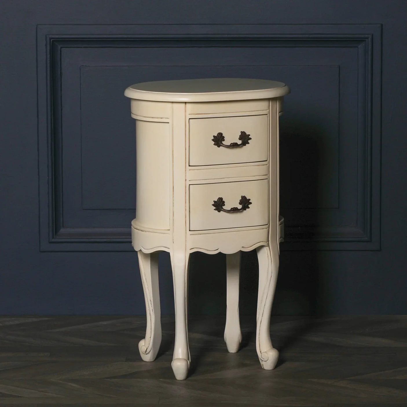 Cream bedside table with two drawers and curved legs against a navy paneled wall