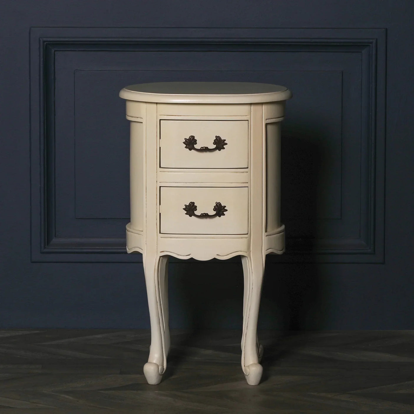 Cream bedside table with two drawers and ornate handles against dark wall