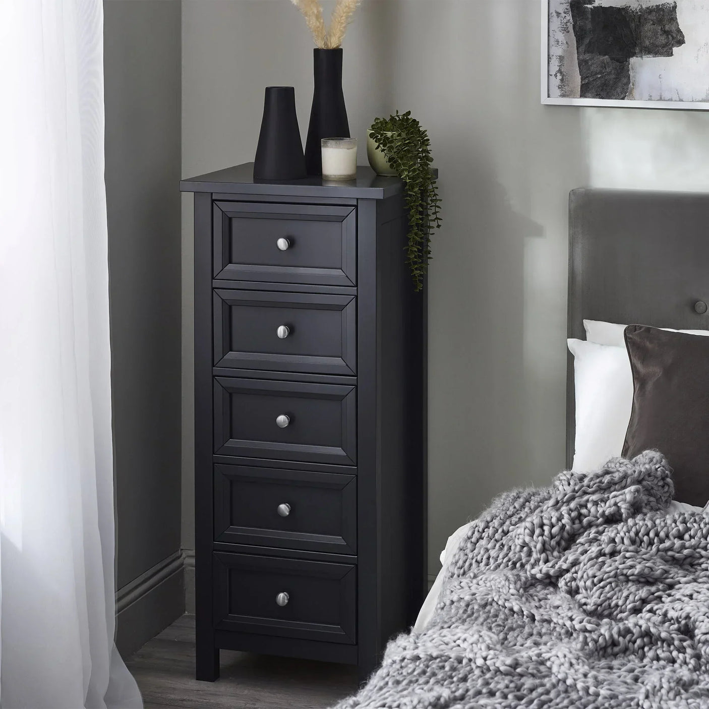 Anthracite 5 drawer tall chest with silver knobs beside a bed in a modern bedroom setting