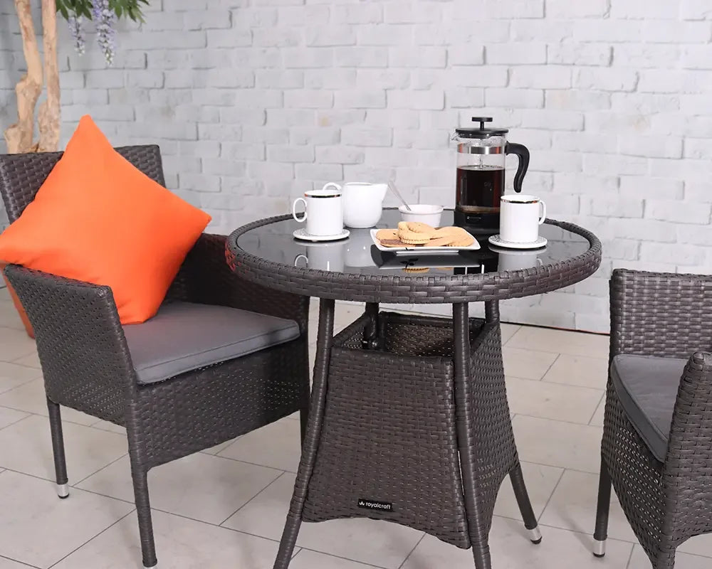 Brown rattan bistro set with round glass table, two chairs, orange cushion and coffee cups