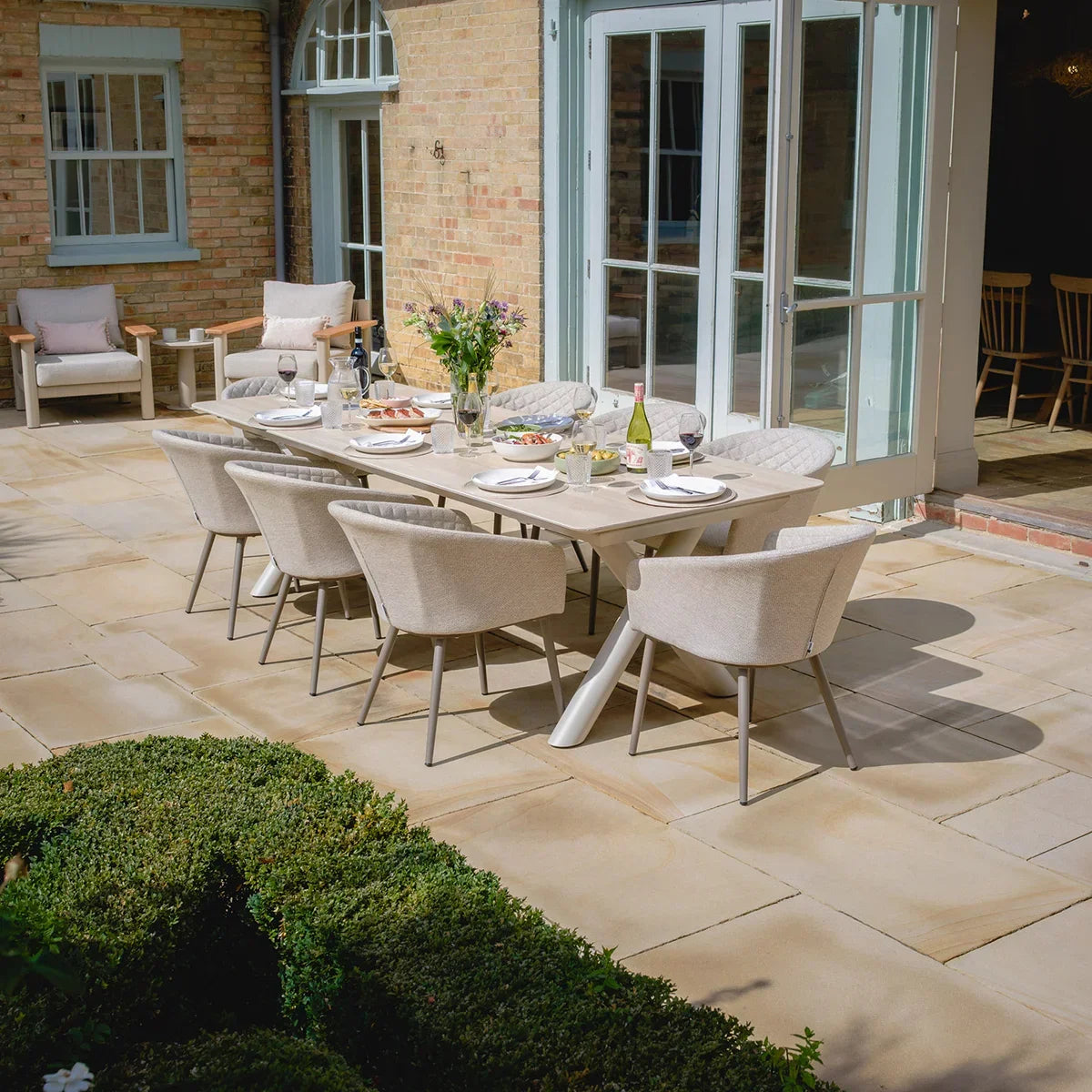 Beige outdoor dining set with eight cushioned chairs on stone patio, table set for lunch