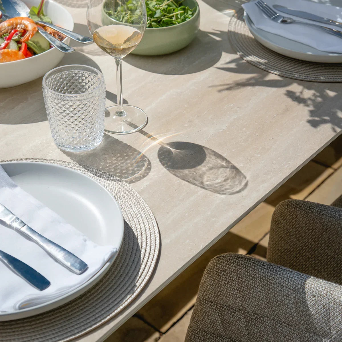 Beige stone-effect dining table set outdoors with plates, glasses, and woven chairs
