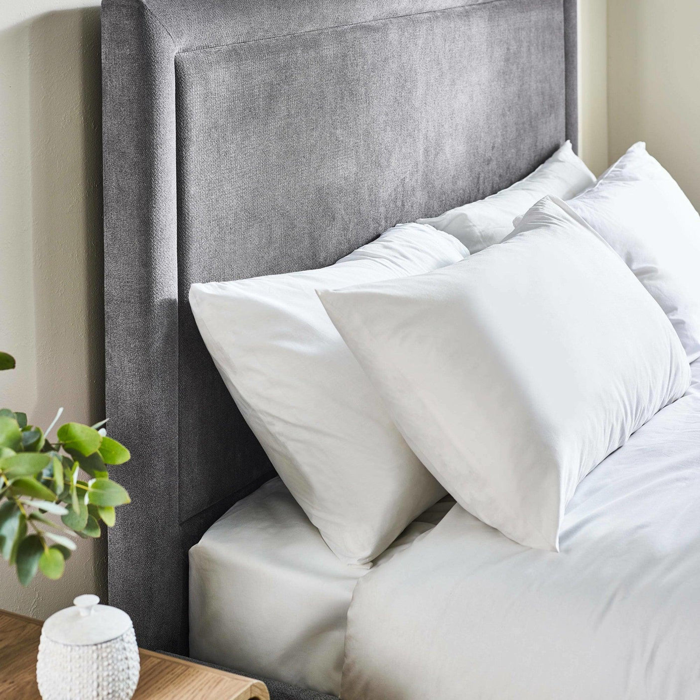 Grey fabric upholstered bed headboard with white bedding and pillows, bedside table visible