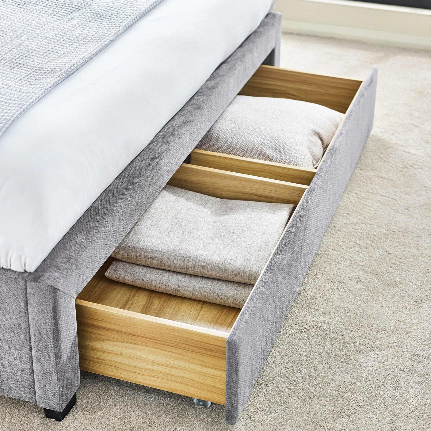 Grey upholstered bed with open wooden storage drawer, bedding and pillows inside