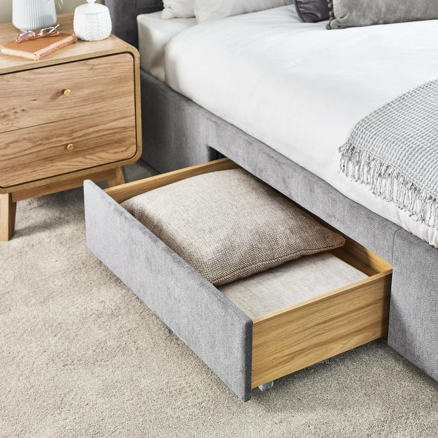Grey fabric storage bed with open drawer containing pillows, next to oak bedside table