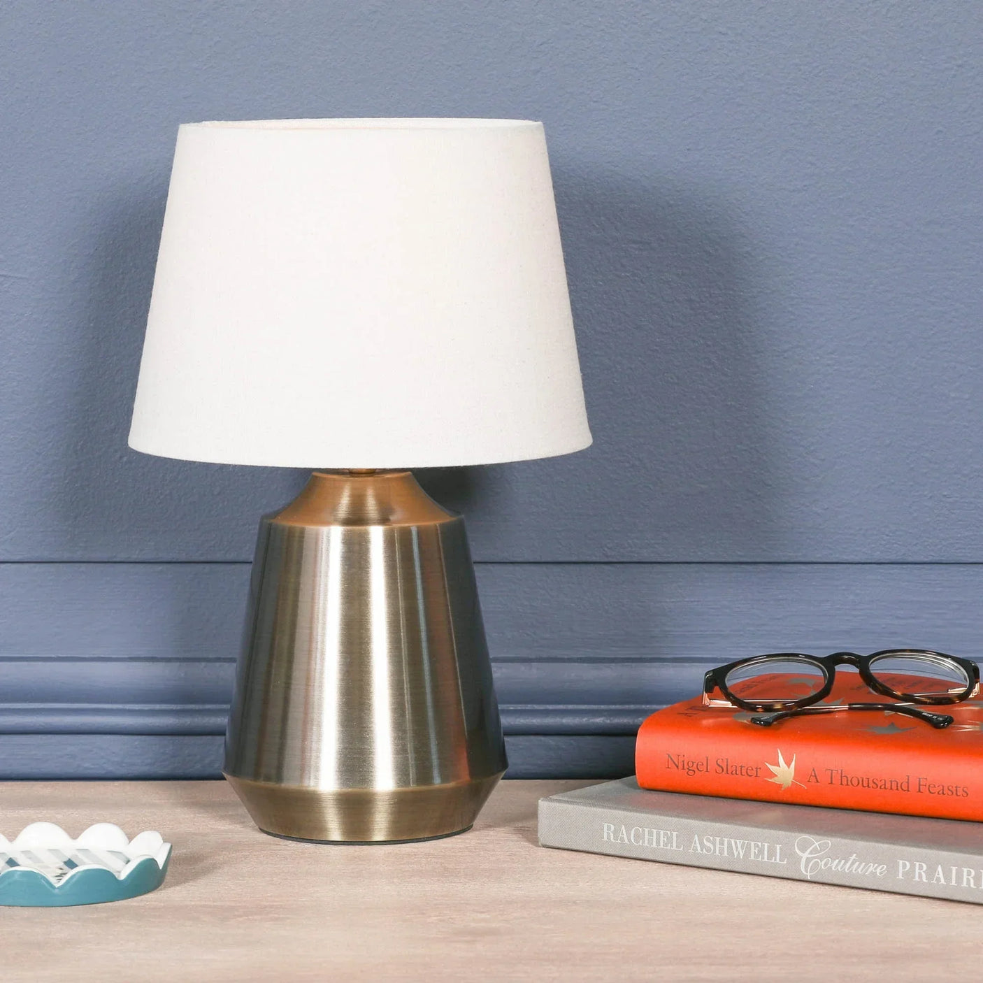 Brushed metal table lamp with white shade on wooden surface beside books and eyeglasses