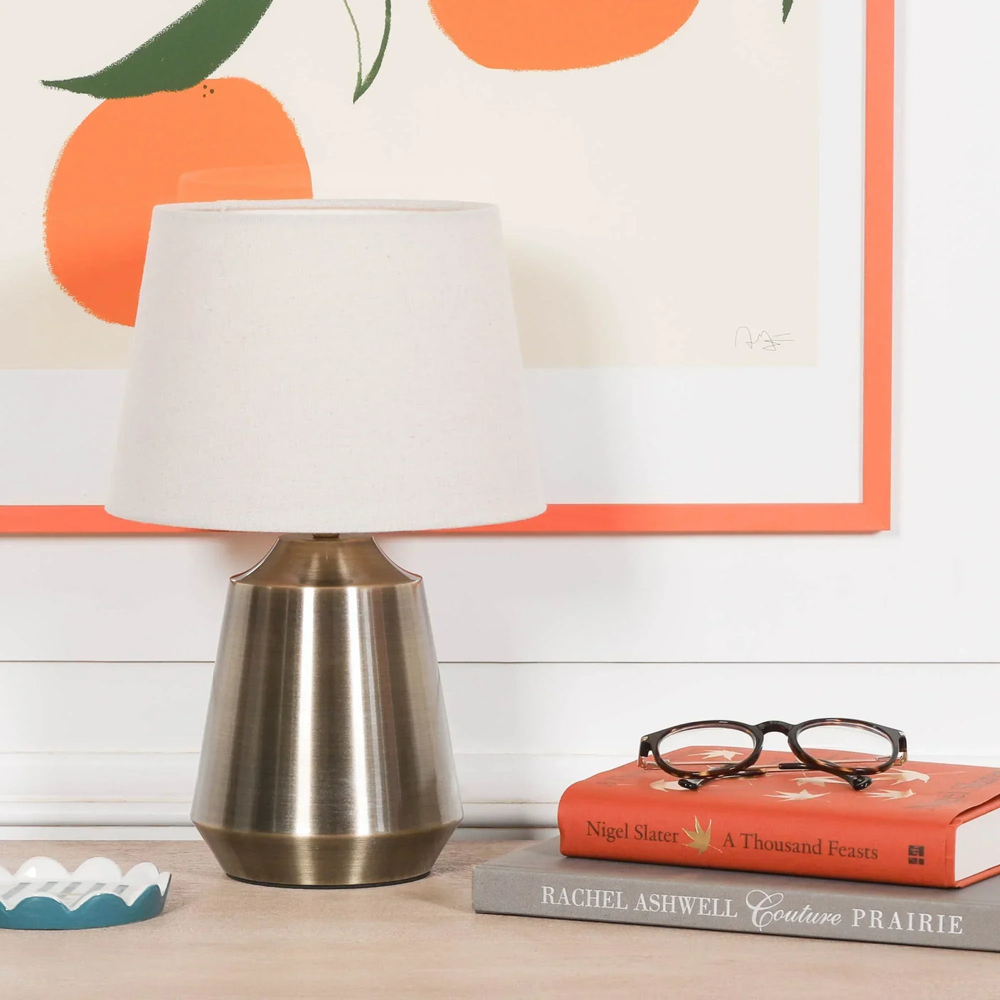 Brushed metal table lamp with white shade on wooden surface beside books and glasses