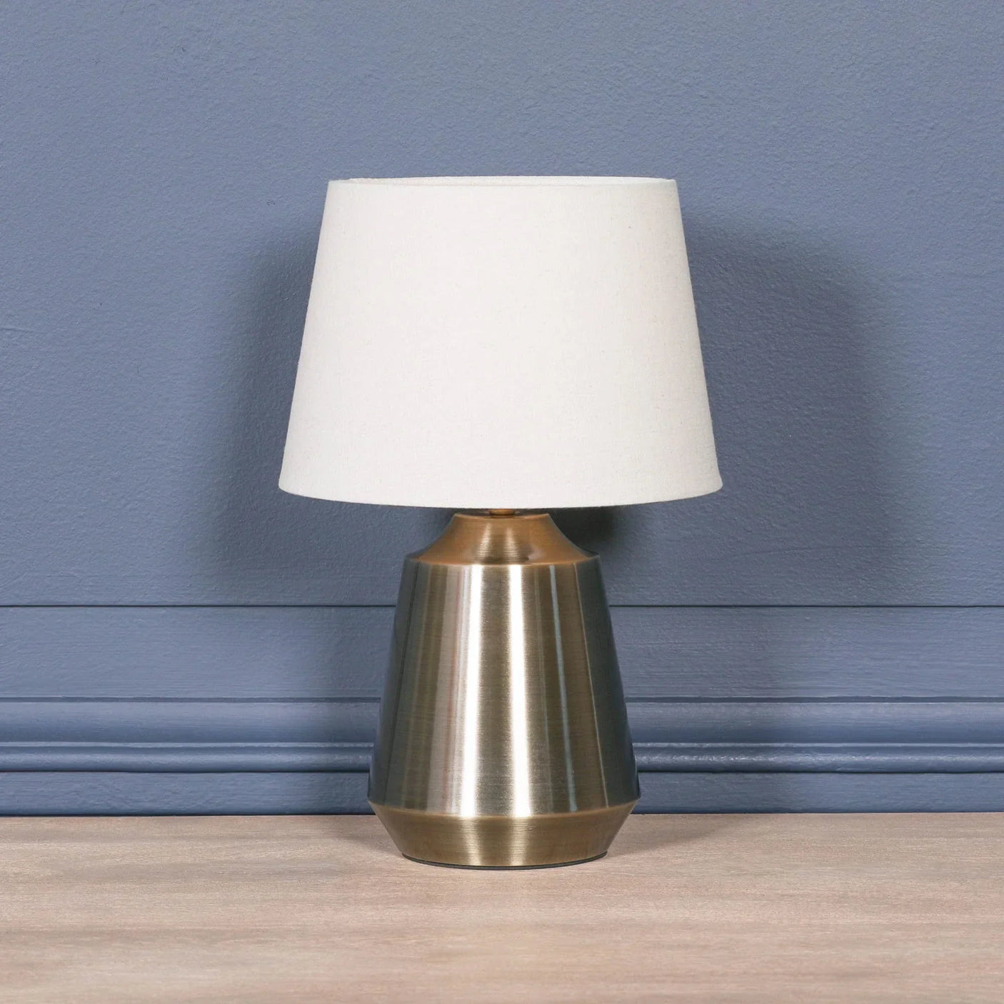 Brushed brass table lamp with white fabric shade on wooden surface against blue wall