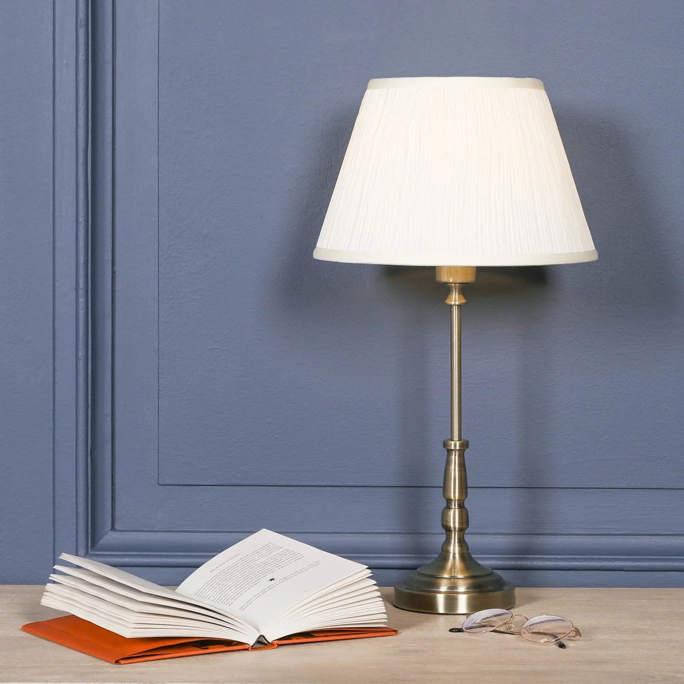 Brass table lamp with cream pleated shade on wooden desk beside open book and glasses