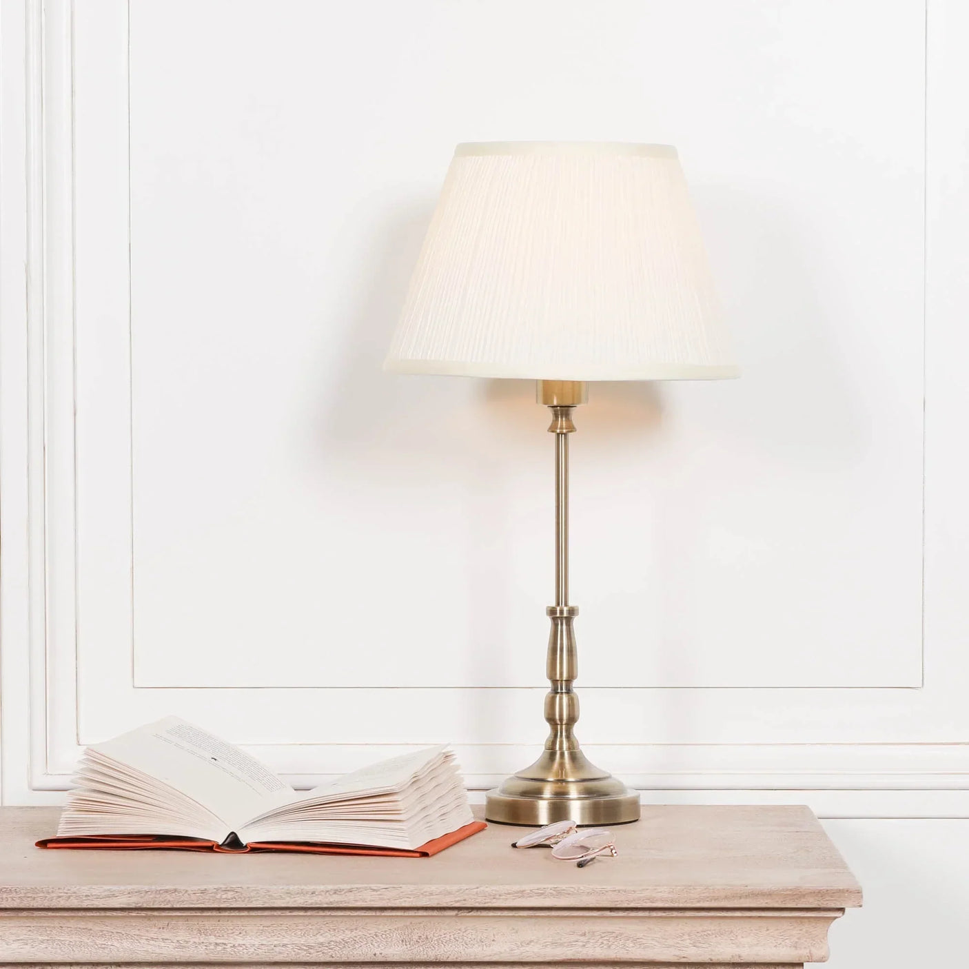 Brass table lamp with cream fabric shade on wooden desk beside open book and glasses