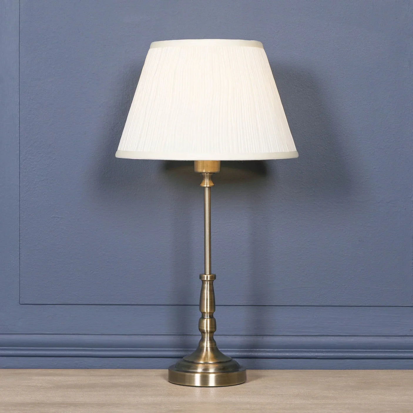 Brass table lamp with white pleated shade against blue wall on wooden surface