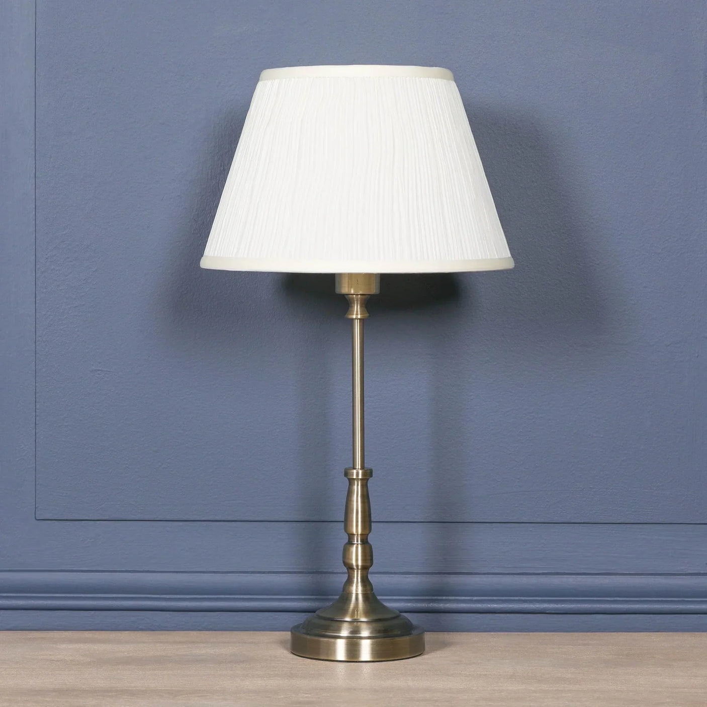 Brass table lamp with white pleated lampshade on wooden surface against blue wall