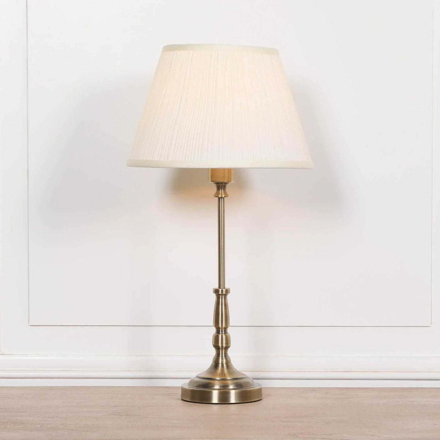 Brass table lamp with cream pleated shade on wooden surface against white panel wall