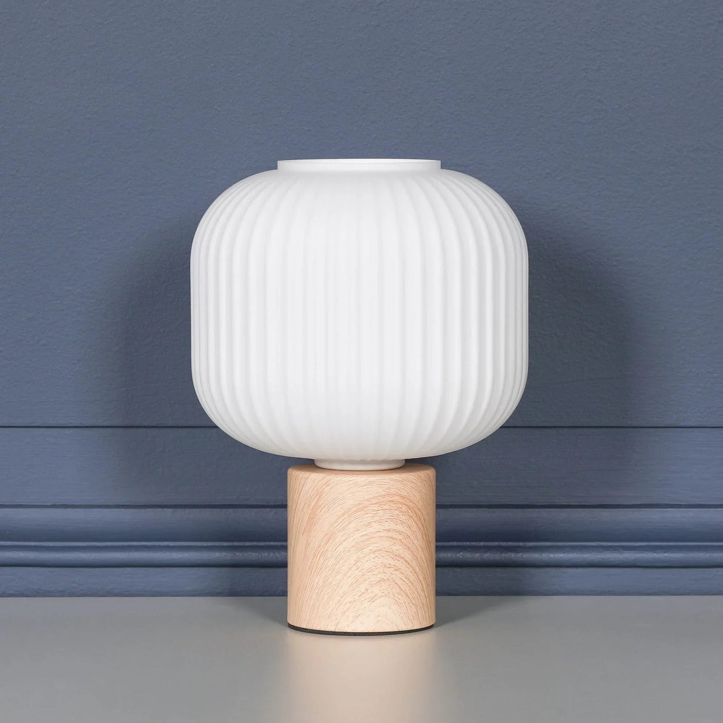 white ribbed glass table lamp with wooden base on grey surface against blue wall