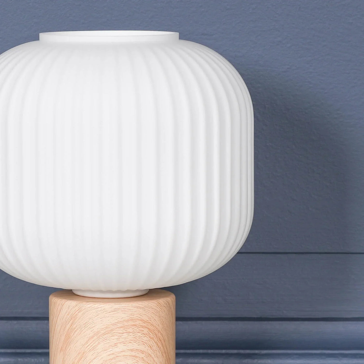 White ribbed glass table lamp with light wood base against blue wall panel