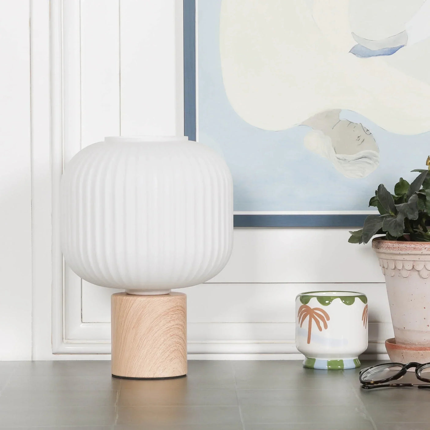 White ribbed table lamp with wooden base beside ceramic plant pot and framed wall art