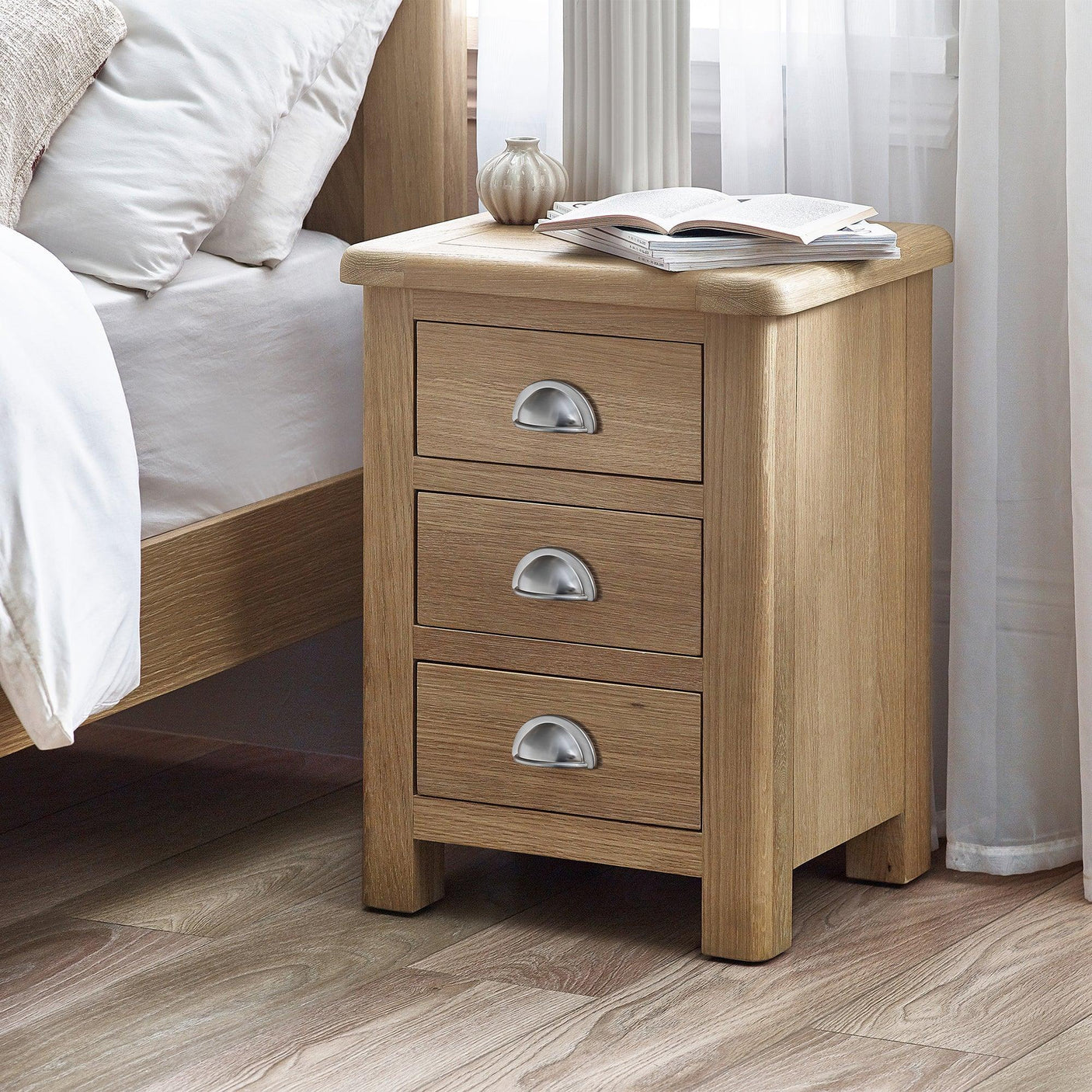 Memphis limed oak 3 drawer bedside table with silver handles beside bed in bright room