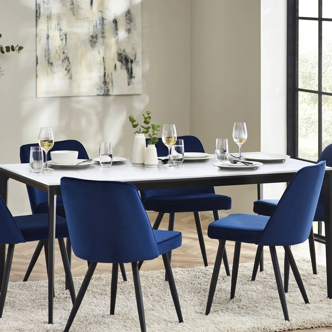 Rectangular stone top dining table with blue chairs and tableware in a bright dining room