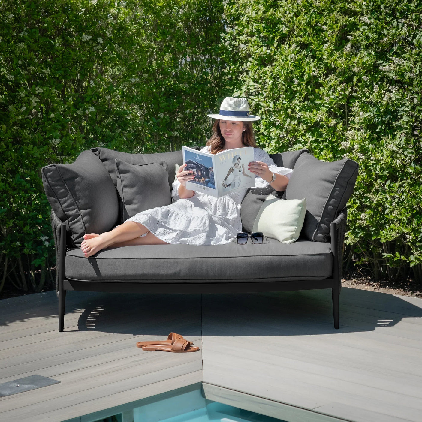 Dark grey outdoor sofa with plush cushions on a patio by pool, person reading magazine