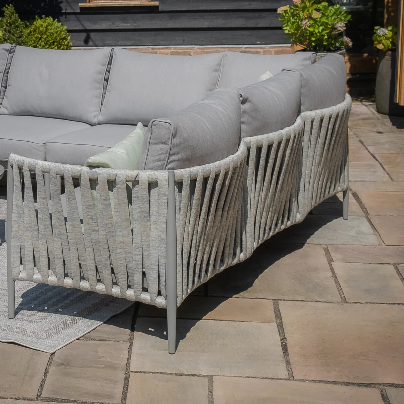 Light grey outdoor corner sofa with rope weave frame and cushions on stone patio