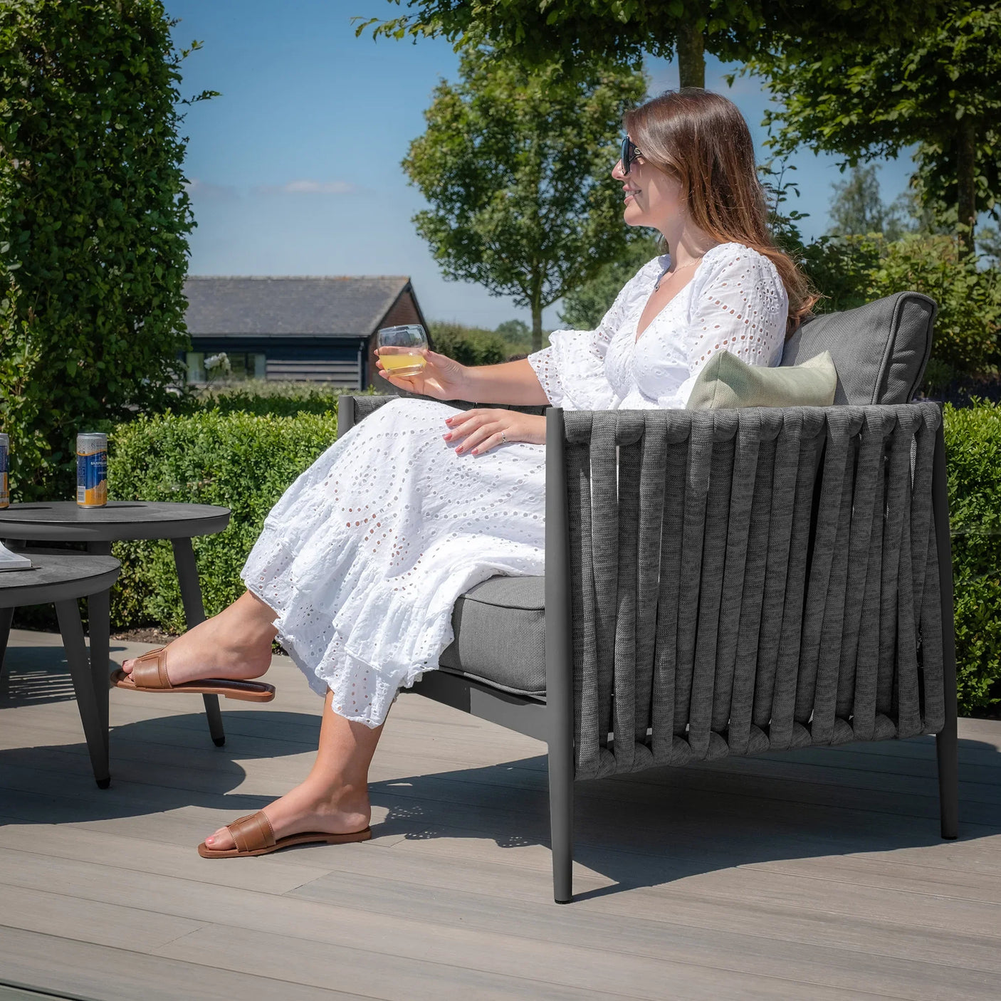 Grey outdoor lounge chair with rope detailing and cushions on patio, woman relaxing in sunlight