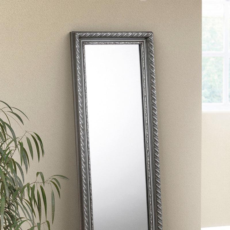 Pewter dress mirror with ornate frame leaning against a beige wall beside houseplant