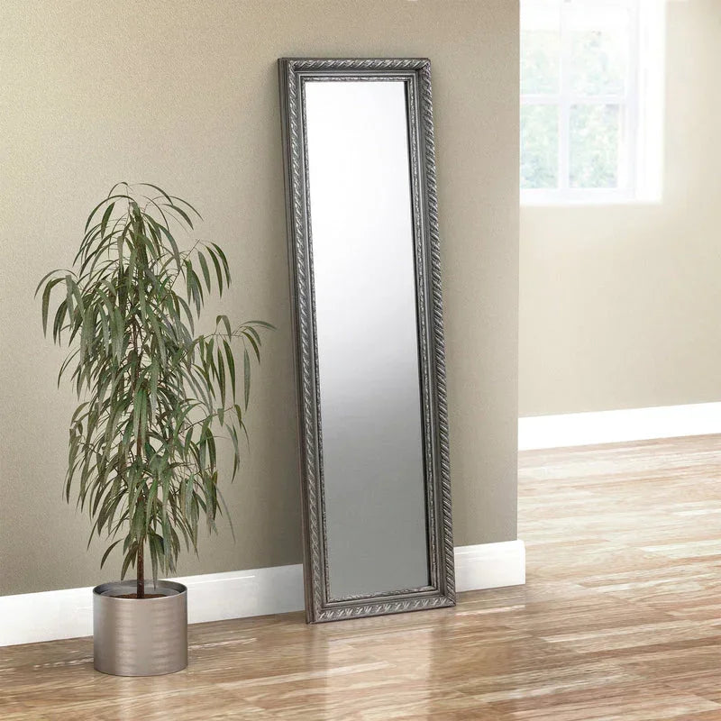Pewter dress mirror with ornate frame leaning against wall beside potted plant on wooden floor