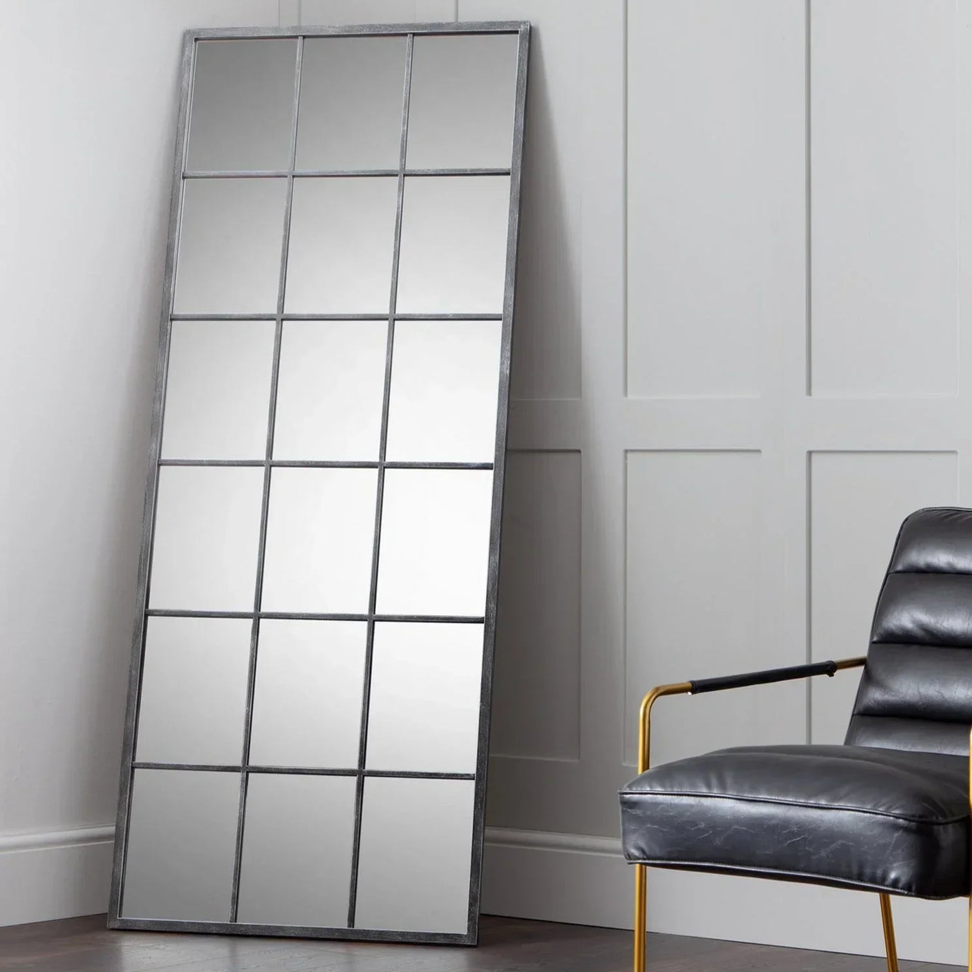 Tall windowpane mirror with black metal frame leaning against panelled wall, armchair beside
