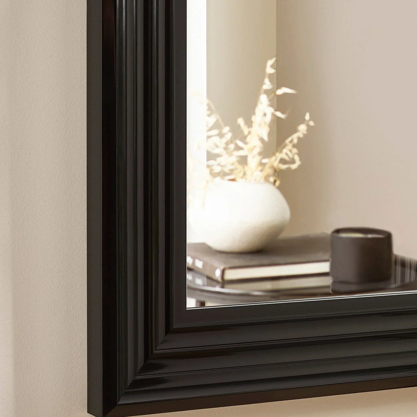 Black wooden mirror frame corner reflecting vase, candle, and stacked books on console table