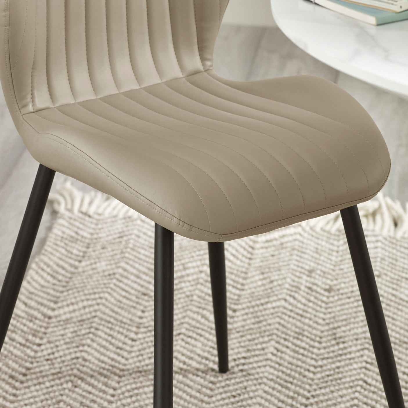 Close-up of beige upholstered dining chair with stitched vertical lines and black metal legs on textured rug.