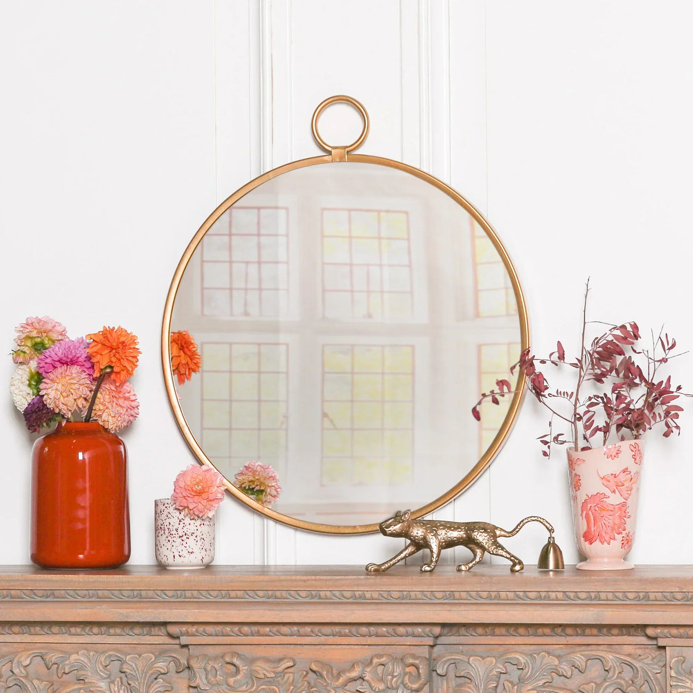 Round gold frame wall mirror on mantel with floral vases and decorative gold cat ornament