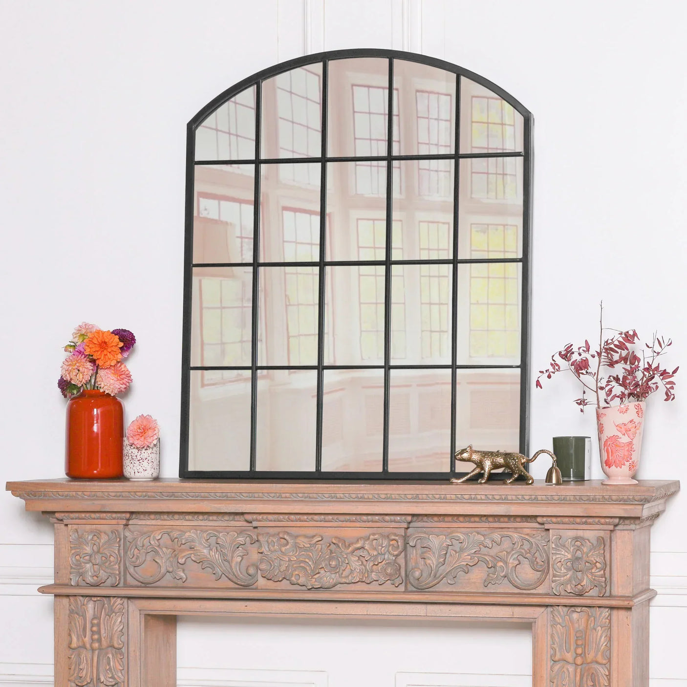 Arched black windowpane mirror on carved wooden mantel with vases and decorative items